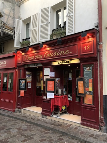 A charming storefront of a red-painted building with the name 'Chez ma Cousine' inscribed in elegant lettering. The facade features large windows with white shutters and a small entrance area displaying several menus and signs, including one advertising hot wine. The lower part of the building is adorned with various posters and bulletin boards, creating a cozy and inviting atmosphere.