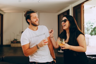 Two people are laughing and enjoying drinks indoors. They appear to be socializing in a casual and relaxed environment, possibly a living room or open space with large glass doors and natural light.