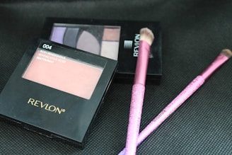 A collection of Revlon makeup products is arranged on a textured black surface, including blush and eyeshadow palettes along with pink makeup brushes. The blush is labeled 'Rosy Rendezvous' and has a matte finish.