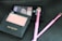 A collection of Revlon makeup products is arranged on a textured black surface, including blush and eyeshadow palettes along with pink makeup brushes. The blush is labeled 'Rosy Rendezvous' and has a matte finish.