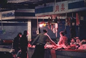 A dimly lit market stall with a vendor and customers. Hanging meats are displayed under a red light, casting a warm glow. People are dressed in dark winter clothing, and one wears a mask, indicating a chilly or urban setting.