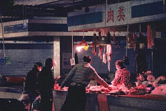 A dimly lit market stall with a vendor and customers. Hanging meats are displayed under a red light, casting a warm glow. People are dressed in dark winter clothing, and one wears a mask, indicating a chilly or urban setting.