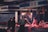 A dimly lit market stall with a vendor and customers. Hanging meats are displayed under a red light, casting a warm glow. People are dressed in dark winter clothing, and one wears a mask, indicating a chilly or urban setting.