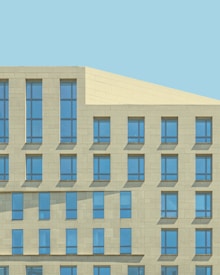 A modern building facade with a minimalist design featuring a grid of rectangular blue-tinted windows set into a light yellow stone exterior. The architecture is characterized by clean, geometric lines and uniform structure.