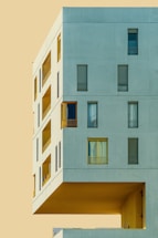 A modern architectural structure with a minimalist design, featuring large rectangular windows and an overhanging section against a flat, pale background.