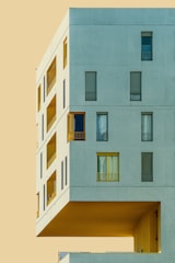 A modern architectural structure with a minimalist design, featuring large rectangular windows and an overhanging section against a flat, pale background.