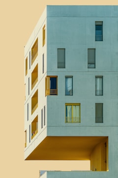 A modern architectural structure with a minimalist design, featuring large rectangular windows and an overhanging section against a flat, pale background.