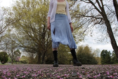 A person wearing a white cardigan, yellow top, blue skirt, and black boots stands on a path covered with scattered pink petals. Trees with budding leaves create a natural backdrop under a cloudy sky.