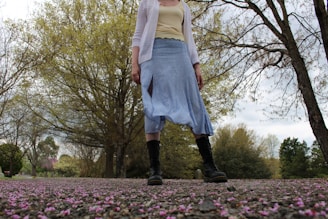 A person wearing a white cardigan, yellow top, blue skirt, and black boots stands on a path covered with scattered pink petals. Trees with budding leaves create a natural backdrop under a cloudy sky.
