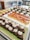 An assortment of desserts is displayed on a countertop. There are small, square pieces of flan on a white rectangular plate, and chocolate brownie squares topped with a dollop of cream on a green rectangular plate. Behind them, there are small cups with a yellow dessert topped with fruit and other small cups with creamy desserts decorated with chocolate bits.