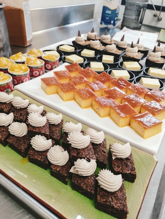 An assortment of desserts is displayed on a countertop. There are small, square pieces of flan on a white rectangular plate, and chocolate brownie squares topped with a dollop of cream on a green rectangular plate. Behind them, there are small cups with a yellow dessert topped with fruit and other small cups with creamy desserts decorated with chocolate bits.