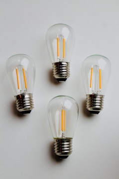 Four clear LED light bulbs are arranged on a plain white surface. The bulbs are turned off, showcasing their distinct filament structure inside each transparent glass casing. The metallic bases of the bulbs stand out noticeably against the white background.