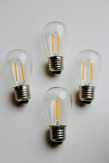 Four clear LED light bulbs are arranged on a plain white surface. The bulbs are turned off, showcasing their distinct filament structure inside each transparent glass casing. The metallic bases of the bulbs stand out noticeably against the white background.