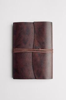 A closed leather notebook with a textured surface and a wraparound strap, lying on a plain white background.