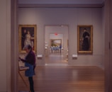 A hallway in an art museum features several framed paintings on the walls, with a woman painting on a canvas in the foreground. The setting is serene and well-lit, with two individuals seated in the background, viewing a large landscape painting on the far wall.