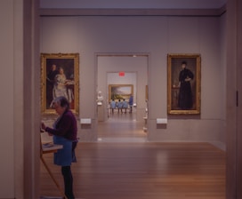 A hallway in an art museum features several framed paintings on the walls, with a woman painting on a canvas in the foreground. The setting is serene and well-lit, with two individuals seated in the background, viewing a large landscape painting on the far wall.