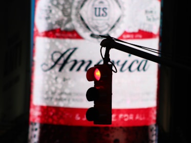A traffic light silhouetted against a large illuminated billboard displaying the word 'America'. The billboard features patriotic colors and imagery, with a prominent red and white color scheme.