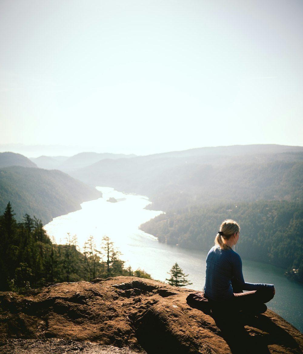 homem na camisa azul de manga comprida sentado na rocha marrom perto do lago durante o dia