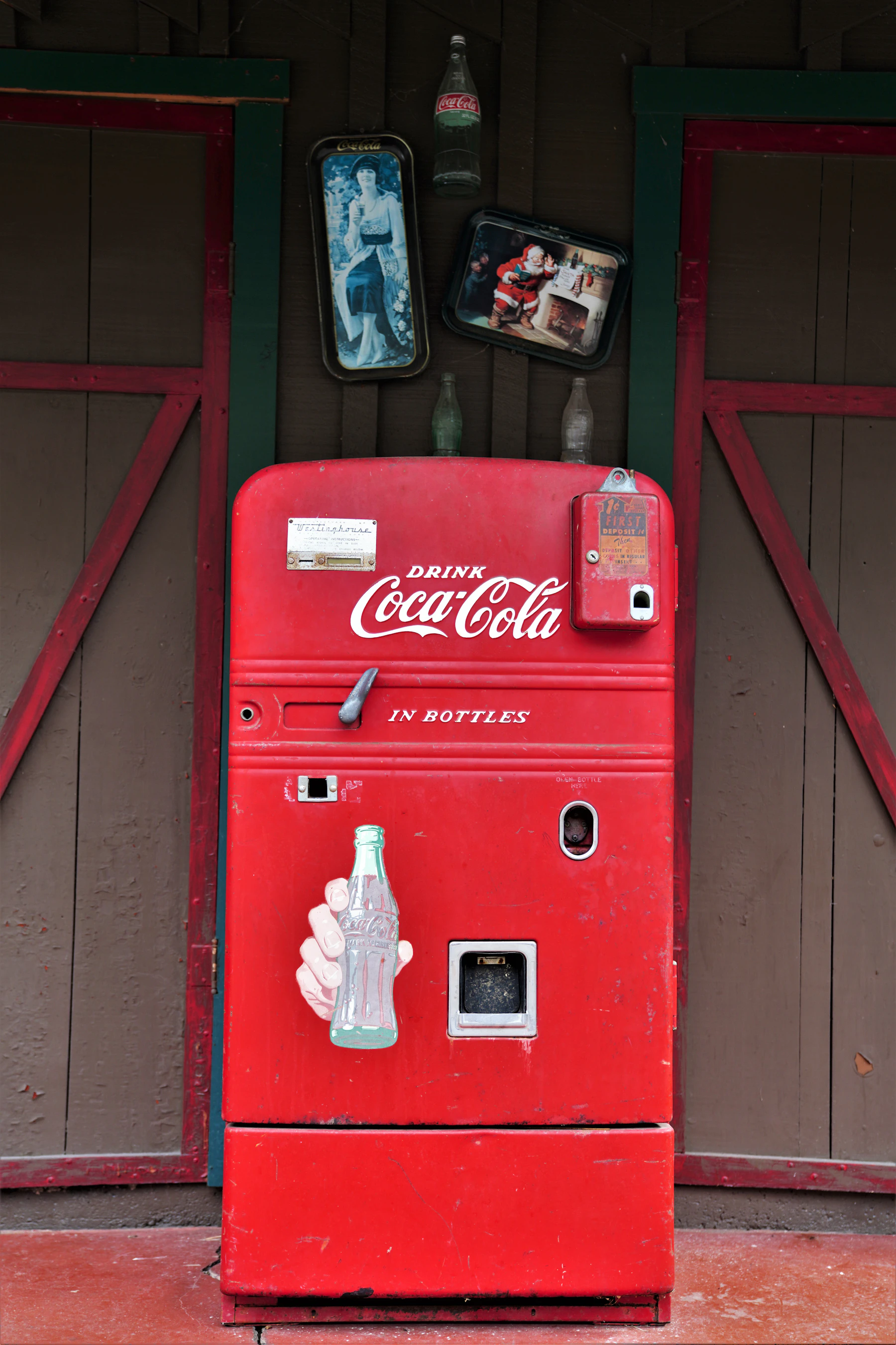 A red upright Coca-Cola bottle vending machine against a barn wall, with two serving trays and three bottles hung above it