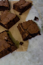 Several square pieces of rich, chocolate brownies are placed on a sheet of parchment paper. The brownies have a crispy top and a dense, moist texture. They are arranged on a marble surface with some crumbs scattered around.