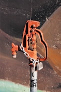 A rusted, orange-colored water valve or hydrant is attached to a gray metallic pipe. The valve has an industrial design and is secured with a metal chain and a lock. The background consists of a textured wall with dark shades of brown and black, accentuated by faint strokes of green at the bottom.