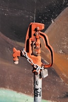 A rusted, orange-colored water valve or hydrant is attached to a gray metallic pipe. The valve has an industrial design and is secured with a metal chain and a lock. The background consists of a textured wall with dark shades of brown and black, accentuated by faint strokes of green at the bottom.