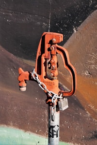 A rusted, orange-colored water valve or hydrant is attached to a gray metallic pipe. The valve has an industrial design and is secured with a metal chain and a lock. The background consists of a textured wall with dark shades of brown and black, accentuated by faint strokes of green at the bottom.