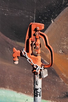 A rusted, orange-colored water valve or hydrant is attached to a gray metallic pipe. The valve has an industrial design and is secured with a metal chain and a lock. The background consists of a textured wall with dark shades of brown and black, accentuated by faint strokes of green at the bottom.