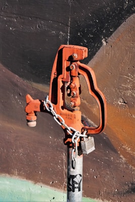 A rusted, orange-colored water valve or hydrant is attached to a gray metallic pipe. The valve has an industrial design and is secured with a metal chain and a lock. The background consists of a textured wall with dark shades of brown and black, accentuated by faint strokes of green at the bottom.
