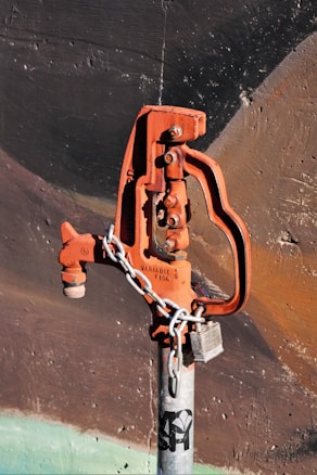 A rusted, orange-colored water valve or hydrant is attached to a gray metallic pipe. The valve has an industrial design and is secured with a metal chain and a lock. The background consists of a textured wall with dark shades of brown and black, accentuated by faint strokes of green at the bottom.