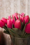 A bouquet of vibrant pink tulips is arranged neatly in a transparent glass vase. The flowers have lush green leaves and are set against a soft, textured background, giving the scene an elegant and serene appearance.