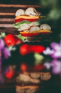 A layered sandwich piled high with various ingredients, including lettuce, tomato, cheese, and bread with a light, rustic texture. The sandwich is set against a blurred background with vibrant colors of red and purple, possibly flowers or vegetables, adding to the aesthetic appeal.