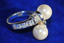 A silver ring adorned with two large pearls and several small rectangular crystals, set against a deep blue background. The pearls are prominently featured, contrasting with the shimmering metal and sparkling stones, giving the piece an elegant appearance.