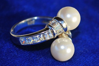 A silver ring adorned with two large pearls and several small rectangular crystals, set against a deep blue background. The pearls are prominently featured, contrasting with the shimmering metal and sparkling stones, giving the piece an elegant appearance.