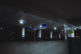 A dimly lit parking garage with signs indicating directions, including 'RIGHT LANE' and 'Arrivals.' The ceiling is supported by white pillars, and the area is enclosed with chain-link fencing. Sparse lighting creates a shadowy atmosphere.