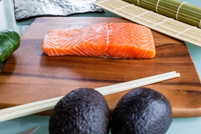 A fresh salmon fillet lies on a wooden cutting board surrounded by sushi-making ingredients. A dark nori seaweed sheet, a bamboo sushi mat, chopsticks, a cucumber, and two avocados are arranged around the board.