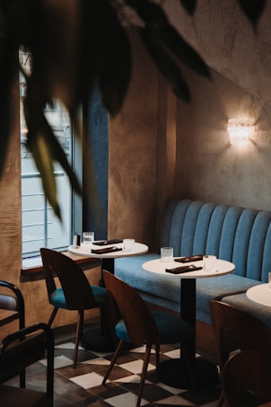 A cozy, dimly lit caf&eacute; interior featuring blue cushioned seating and wooden chairs. Two round tables are set with glassware and napkins, positioned near a warmly illuminated wall sconce. Natural light streams in through a window, casting soft shadows across the checkered floor tiles.