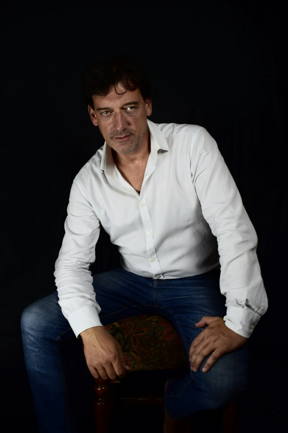 man in white dress shirt and blue denim jeans sitting on brown wooden stool