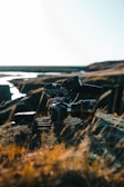 A professional camera setup, equipped with a large lens and mounted on a stabilizer or tripod, rests on rocky terrain with sparse grass. The background shows a river or stream winding through a barren landscape under a bright, clear sky.