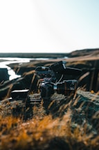 A professional camera setup, equipped with a large lens and mounted on a stabilizer or tripod, rests on rocky terrain with sparse grass. The background shows a river or stream winding through a barren landscape under a bright, clear sky.