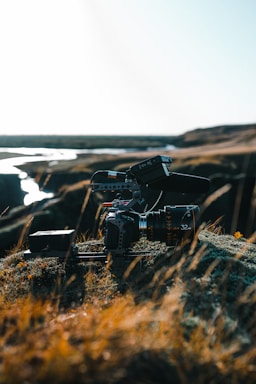A professional camera setup, equipped with a large lens and mounted on a stabilizer or tripod, rests on rocky terrain with sparse grass. The background shows a river or stream winding through a barren landscape under a bright, clear sky.
