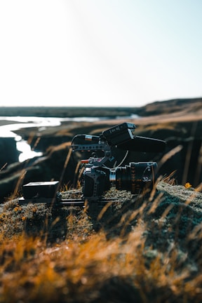 A professional camera setup, equipped with a large lens and mounted on a stabilizer or tripod, rests on rocky terrain with sparse grass. The background shows a river or stream winding through a barren landscape under a bright, clear sky.