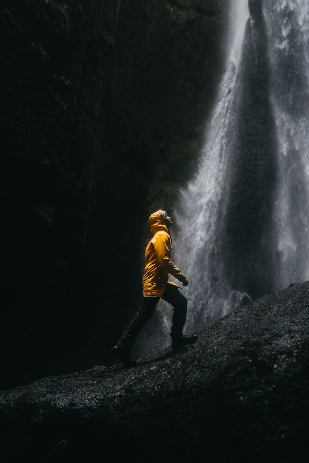 woman in yellow jacket and black pants standing on rock in front of waterfalls