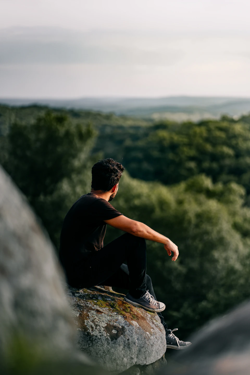 Man sitting on large rock formation, dressed casually, lost in thought outdoors