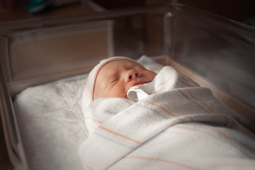 A newborn baby wrapped in a soft blanket rests peacefully in a hospital-grade bassinet. The lighting is soft and warm, creating a serene atmosphere.
