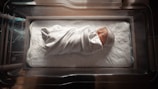 A newborn baby wrapped in a white blanket with subtle colored stripes, lying peacefully in a hospital bassinet. The baby is wearing a small knitted cap and is placed on a thick, soft mattress that lines the bassinet. Natural light softly illuminates the scene, casting gentle shadows around the baby.