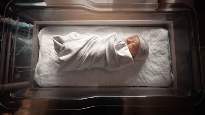 A newborn baby wrapped in a white blanket with subtle colored stripes, lying peacefully in a hospital bassinet. The baby is wearing a small knitted cap and is placed on a thick, soft mattress that lines the bassinet. Natural light softly illuminates the scene, casting gentle shadows around the baby.