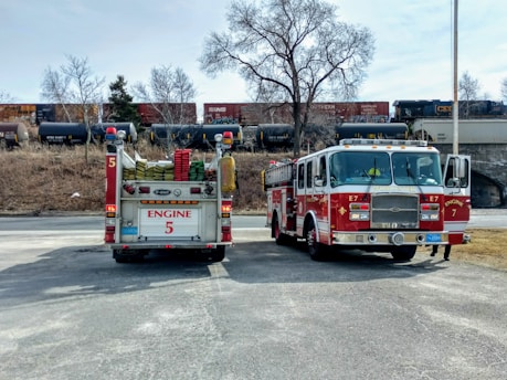 Two fire trucks are parked on a paved surface with a backdrop of trees, grass, and a freight train on elevated tracks. One fire truck shows the back view, displaying hoses and the label 'Engine 5', while the other truck displays a front view with the label 'Engine 7' and 'Worcester'. The scene includes a clear sky and a railroad bridge.