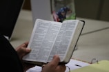 A person is holding an open book, which appears to be a religious text or Bible. The text is in Spanish and is printed in two columns. The person's hands are visible, and there is a Captain America-themed mug and a binder on the table.