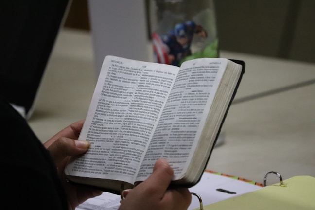 A person is holding an open book, which appears to be a religious text or Bible. The text is in Spanish and is printed in two columns. The person's hands are visible, and there is a Captain America-themed mug and a binder on the table.